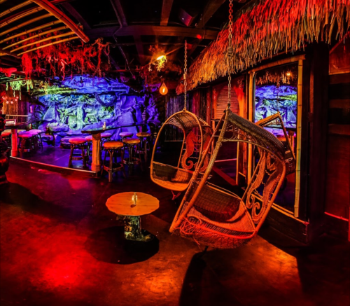 A vibrant, tropical-themed bar interior with hanging chairs, colorful lighting, and a rustic wooden decor.