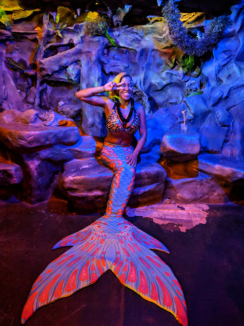 A person in a colorful mermaid costume poses in a vibrant underwater-themed setting with rocks and plants.