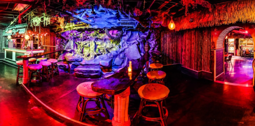 A vibrant bar scene with colorful lighting, rock formations, and wooden stools in a tropical-themed setting.