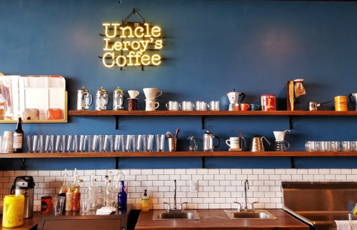 A cozy coffee shop interior featuring a neon sign, shelves with mugs, and a clean countertop with glassware.