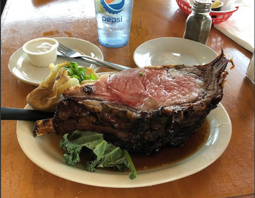 A large prime rib steak served with a baked potato, green onions, and a side of creamy sauce on a plate.