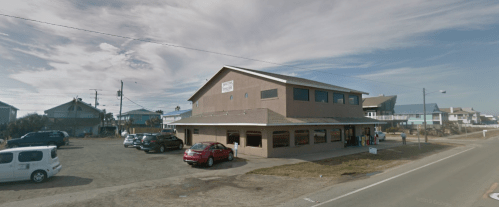 A two-story building with a sign, surrounded by parked cars and a clear sky, near a coastal area.