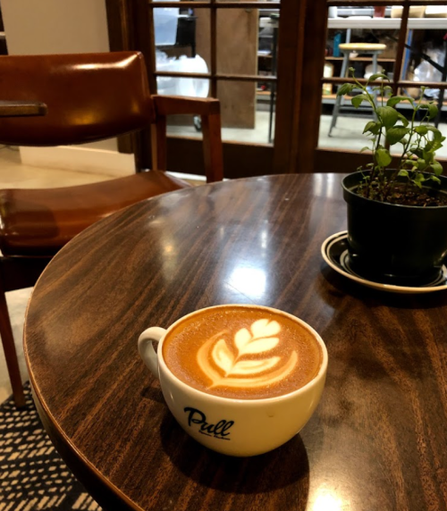 A latte with a leaf design sits on a wooden table next to a small potted plant and a wooden chair in a cozy café.