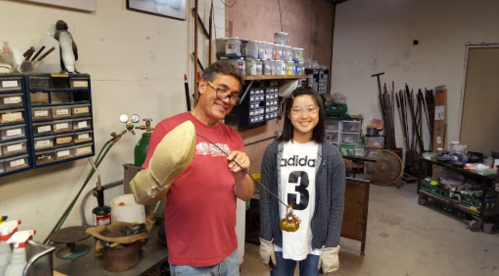 A man and a girl smile in a workshop, holding glassblowing tools and wearing gloves, surrounded by equipment.