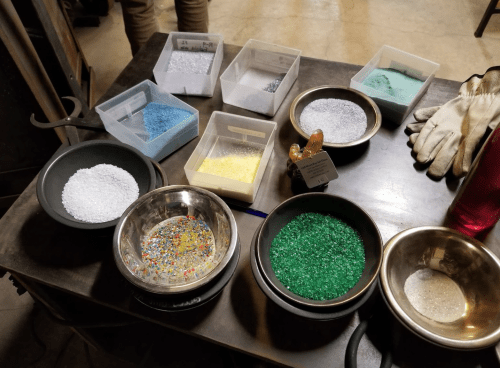A table with various bowls and containers filled with colorful glass granules and powders, alongside gloves and a water bottle.