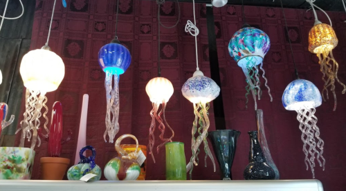 Colorful glass jellyfish-shaped pendant lights hanging against a textured maroon wall, with various vases below.