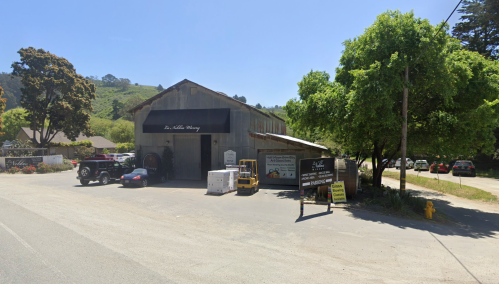 A rustic winery building with a sign, surrounded by trees and parked vehicles on a sunny day.