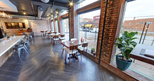 A modern restaurant interior with wooden tables, chairs, and large windows showcasing a street view.