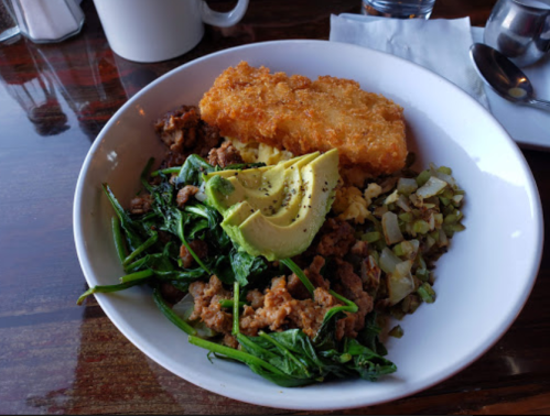 A bowl of scrambled eggs with sausage, sautéed greens, avocado slices, and a crispy hash brown on top.