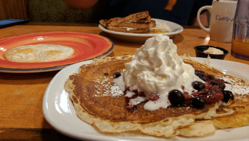 A plate of pancakes topped with whipped cream and berries, with a side of eggs and toast in the background.