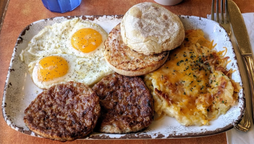 A breakfast plate featuring two fried eggs, sausage patties, hash browns, and toasted English muffins.