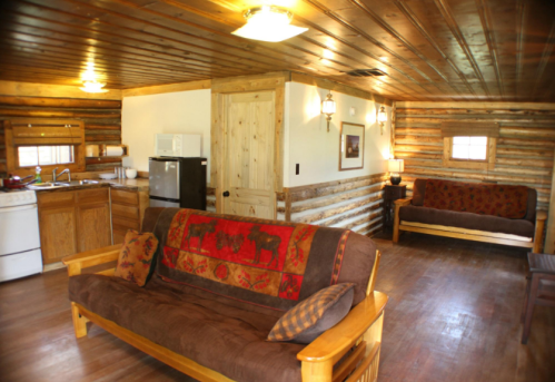 Cozy cabin interior with wooden walls, a kitchen area, and comfortable seating. Warm lighting creates a welcoming atmosphere.