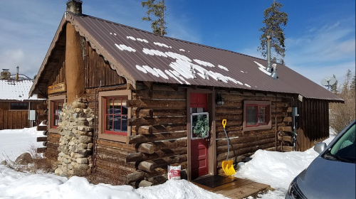 A rustic log cabin with a snow-covered roof, red door, and stone accents, surrounded by snow and trees.