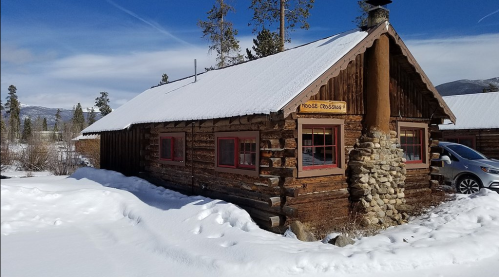 A rustic log cabin with a stone chimney, surrounded by snow and pine trees under a clear blue sky.