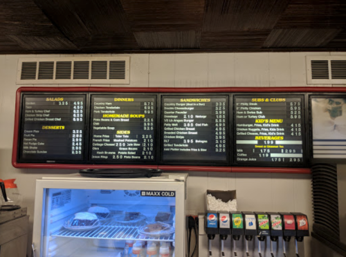 Menu board displaying salads, dinners, sandwiches, subs, clubs, homemade soups, desserts, and beverages with prices.