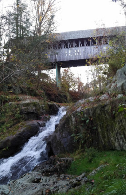 A covered bridge stands above a rocky stream, surrounded by trees and autumn foliage.