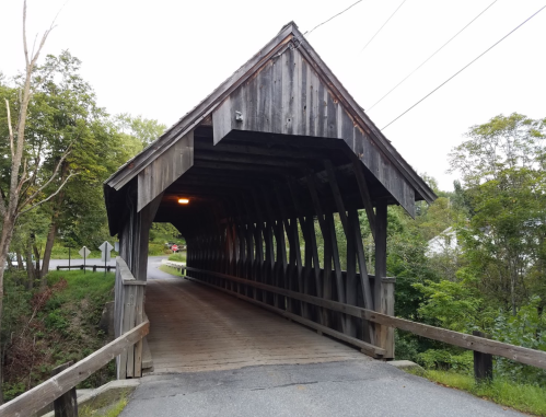 A wooden covered bridge with a sloped roof, surrounded by trees and a road leading into the structure.