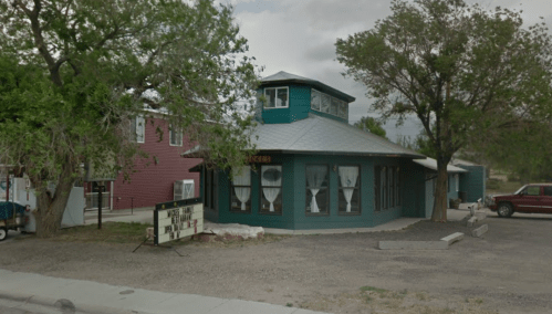 A teal building with a unique roof design, surrounded by trees and gravel, featuring a sign in front.