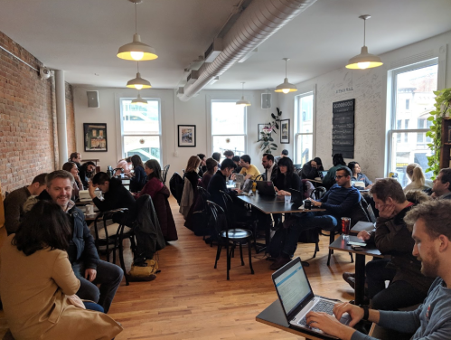 A busy café with people working and chatting, featuring wooden floors and large windows.