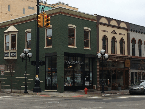A green corner bookstore with large windows, surrounded by historic buildings and street lamps on a cloudy day.