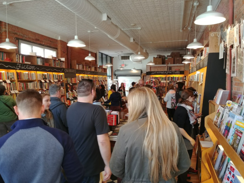 A busy bookstore interior with people browsing bookshelves filled with various genres. Bright and inviting atmosphere.