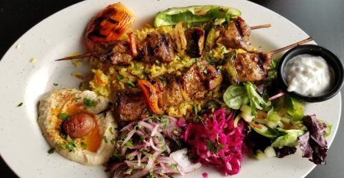 A plate of grilled skewers with rice, salad, hummus, pickled vegetables, and a side of yogurt sauce.