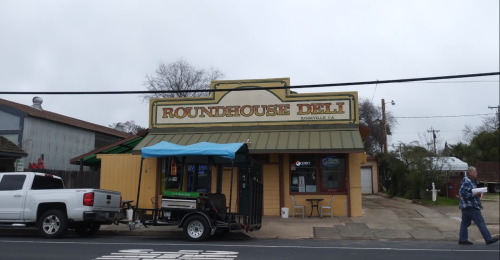 A yellow deli building with a green roof, featuring a sign that reads "Roundhouse Deli" in a small town.