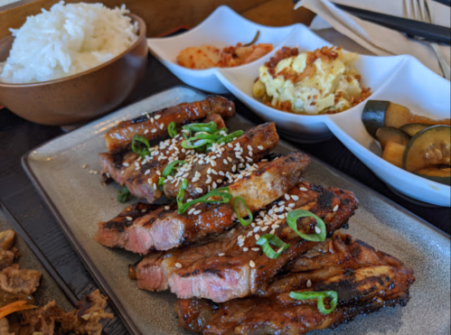 A plate of grilled ribs topped with sesame seeds and green onions, served with rice, kimchi, and side dishes.