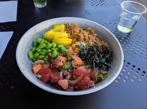 A bowl of colorful poke with salmon, tuna, edamame, mango, seaweed, and rice on a black table.