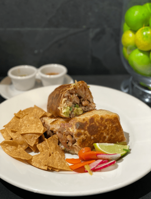 A plate with a halved burrito, tortilla chips, pickled vegetables, lime wedges, and two small dipping sauces.