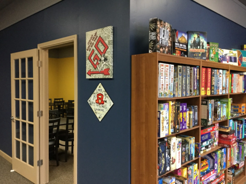 A blue wall with a board game shelf and a doorway leading to a room with chairs and tables.