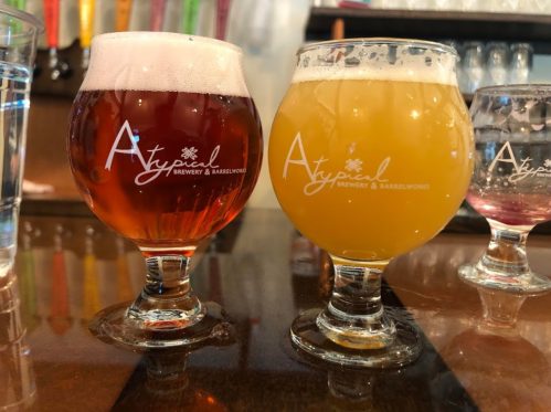 Two craft beer glasses on a bar: one with a reddish hue and the other a hazy yellow, both branded with "Atypical Brewery."