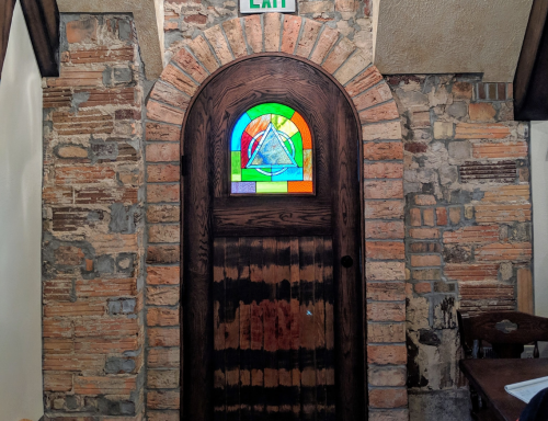 A wooden door with a colorful stained glass window, set in a stone wall, marked with an "EXIT" sign above.