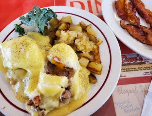 A plate of eggs Benedict with sausage, hollandaise sauce, diced potatoes, and a side of crispy bacon.