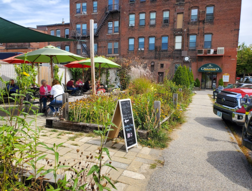 A vibrant outdoor café with colorful umbrellas, surrounded by greenery and flowers, next to a brick building.