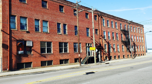 A red brick building with multiple windows and a fire escape, located along a street under a clear blue sky.