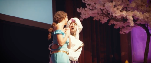 A woman in a blue dress and a man in a white outfit perform together on stage, surrounded by cherry blossom decorations.