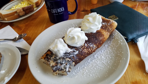 A large pancake roll topped with whipped cream and powdered sugar, served on a white plate with a coffee mug in the background.
