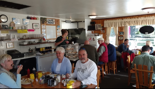 A cozy café scene with patrons enjoying meals, a server at the counter, and a warm, inviting atmosphere.