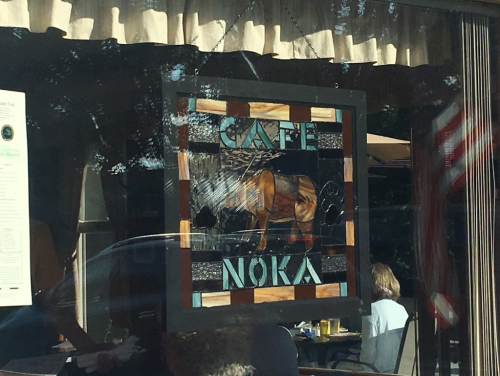 Stained glass sign reading "Café Noka" with a horse design, seen through a café window.