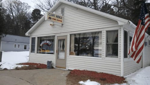 A cozy white cafe with a sign reading "Cafe Noka" and an American flag, surrounded by snow and trees.