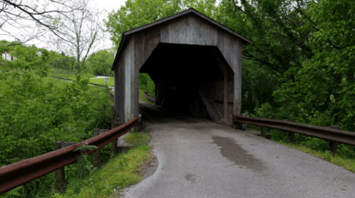 A wooden covered bridge over a road, surrounded by lush green trees and grass.