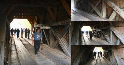 People walking through a wooden covered bridge with rustic beams and a weathered floor, surrounded by autumn foliage.