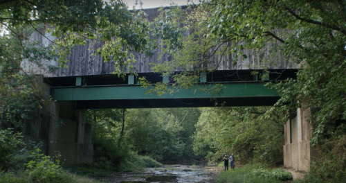 A green bridge spans a shallow creek, surrounded by lush greenery and trees, with two people standing nearby.