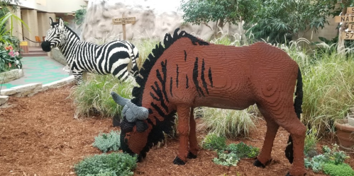 A life-sized LEGO sculpture of a brown horse and a zebra in a lush indoor setting with plants and rocks.