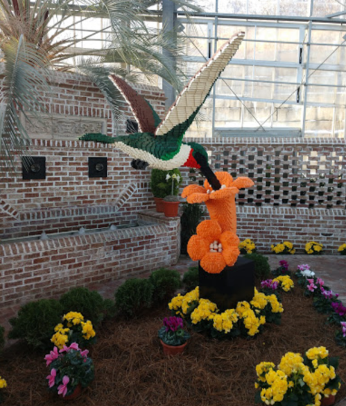A colorful sculpture of a hummingbird hovering over an orange flower, surrounded by vibrant blooms in a greenhouse.