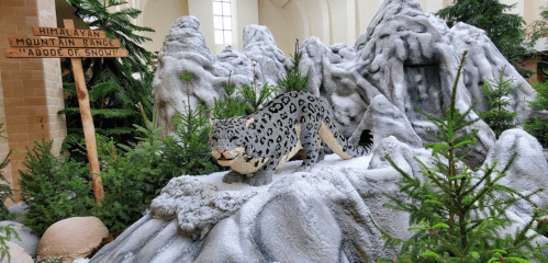 A LEGO sculpture of a snow leopard on a snowy mountain landscape, surrounded by greenery and a sign about the Himalayan range.