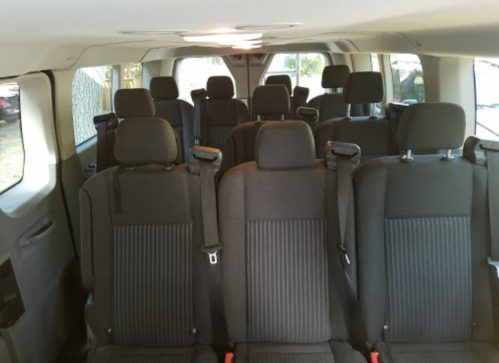 Interior of a spacious van with multiple rows of black fabric seats, all facing forward and equipped with seatbelts.