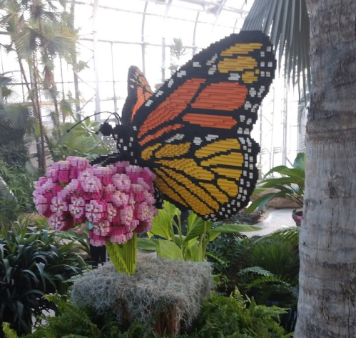 A large butterfly made of colorful blocks perched on a pink flower, surrounded by lush greenery in a greenhouse.