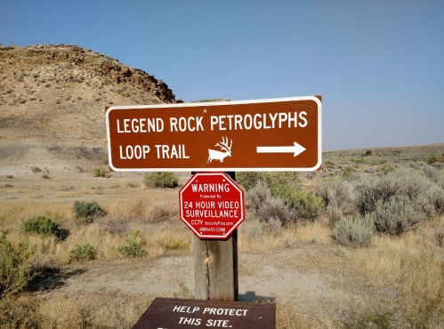 Sign for Legend Rock Petroglyphs Loop Trail with a warning about video surveillance, surrounded by rocky terrain and shrubs.